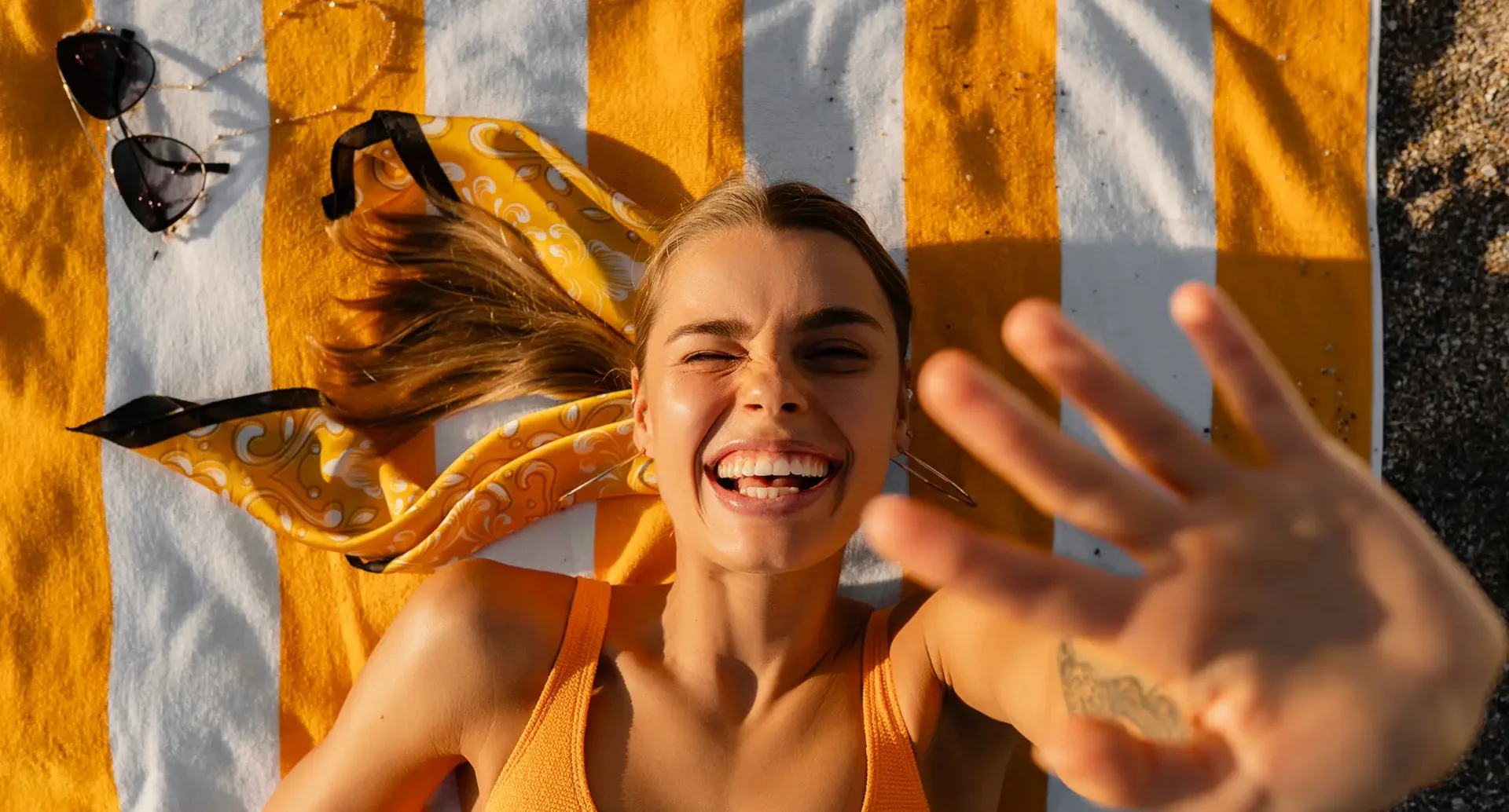Nahaufnahme einer Person auf einem gelb-weiß gestreiften Strandtuch mit ausgestreckter Hand beim Selfie, mit Sonnenbrille und gelbem Haarband daneben